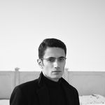 Black and white portrait of a serious man in a coat standing outdoors against a wall.